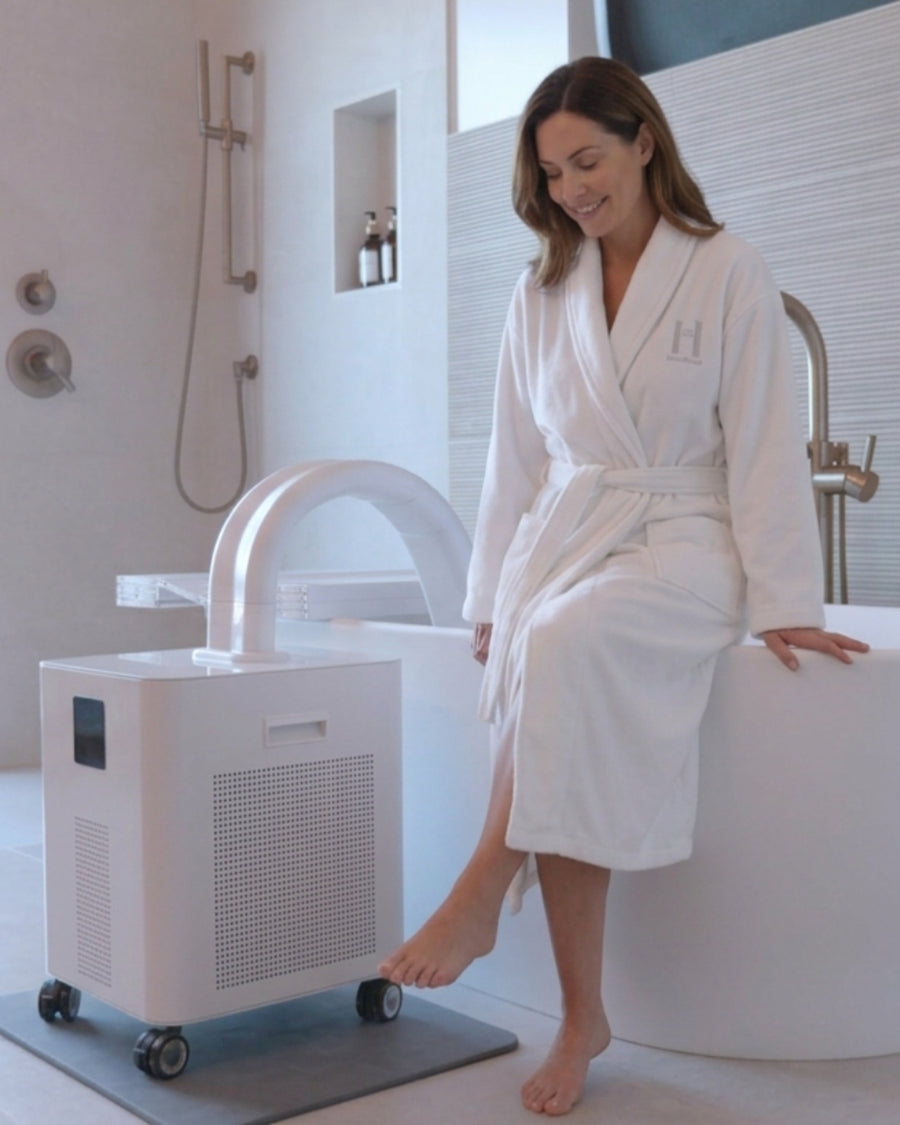 A HomePlunge owner in the HomePlunge robe sitting on the edge of her bathtub, the chiller beside her with the hose-arm over the rim and the Bath Stone on the floor